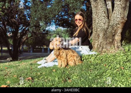 A woman sits under a tree, smiling as she shares a moment of joy with her cocker spaniel. The sun shines down on them in a peaceful park setting, surr Stock Photo