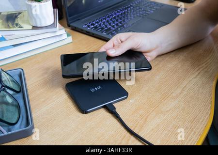 Girl hand placing smartphone on wireless charging pad on working table with notebook, eyeglasses, and books Stock Photo