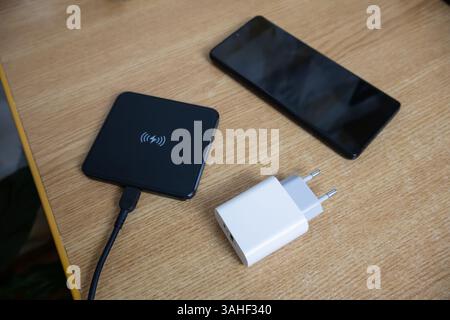 White charger used to charge empty battery, black wireless charging pad and smartphone placed on the table, top view Stock Photo