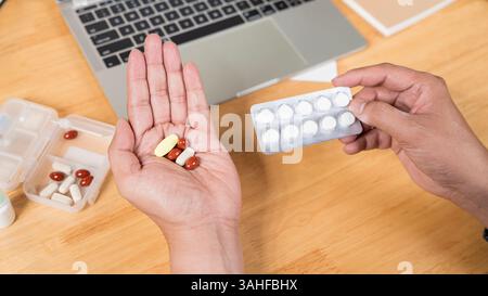 Close-up of a male hand holding a pill bottle pouring medication into his hand. Young man taking pills from pillbox while using laptop at home. Stock Photo