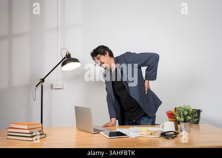 Young businessman having acute back pain, massaging aching muscles, sitting at desk in modern office, copy space. Millennial guy having health problem Stock Photo