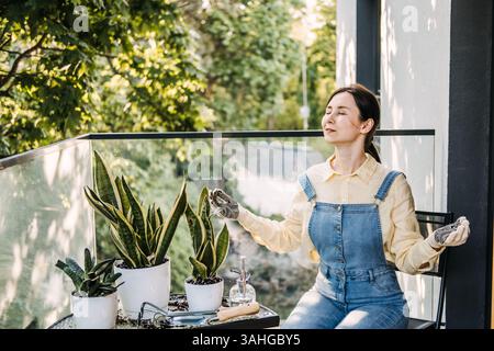 Woman meditating on a balcony with raised arms among potted plants in an urban garden space. Balcony gardening, nature in the city, plant therapy Stock Photo