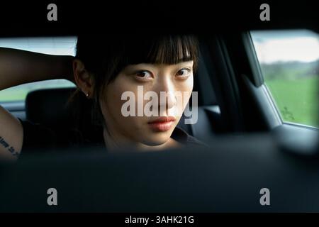 Woman with serious expression sits in car, holding hair back, looking at camera through rearview mirror. Brandenburg, Germany Stock Photo