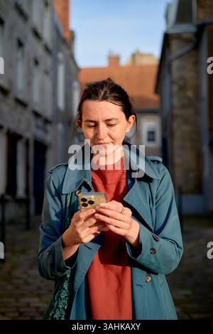 Woman in blue coat using smartphone on a cobblestone street with old buildings. Normandy, France Stock Photo