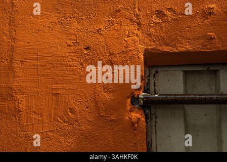 Close-up of a vivid orange wall with a small window and metal security bar, showing thick textured paint Stock Photo