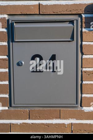 A modern gray mailbox with the number 24 is embedded in a brick wall, featuring a lock and a mail slot for secure postal deliveries. Stock Photo