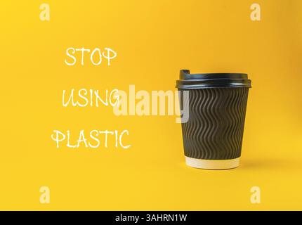 Black coffee cup with a white lid sits on a yellow background. The words Stop using plastic are written below the cup. earth day holiday concept Stock Photo