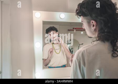 Young artist with a mullet haircut and a white t-shirt looking at himself in the mirror in his studio, adjusting his glasses and touching his neck Stock Photo