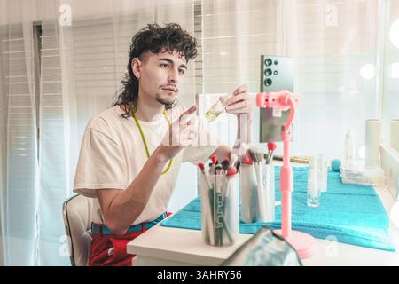 Young latin male beauty influencer showing skincare product while recording video for social media using smartphone at home, promoting cosmetics to hi Stock Photo