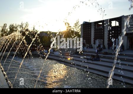 Water jets and drops of a fountain into the pool viewed in back light at sunset on blue sunny sky background. Building and staircase in the background Stock Photo