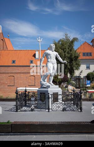 A statue of a man holding a spear is in front of a red brick building. The statue is surrounded by a fence and a bench Stock Photo
