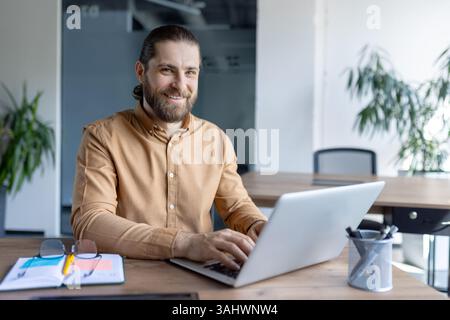 A bearded man smiles at the camera while working on a laptop. The office environment shows the focus on productivity and work. Stock Photo