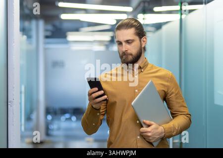 A man with a beard checks his phone while holding a laptop, walking down a hallway. The business professional is likely checking emails or messages. Stock Photo
