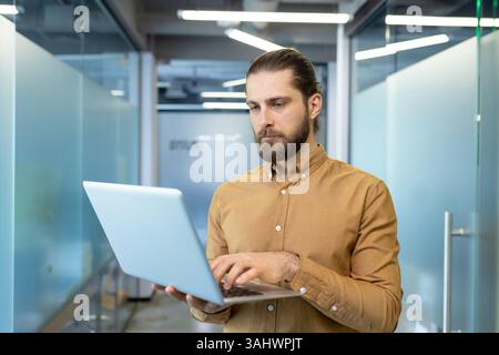 A professional man, focused on his work, types on a laptop in a modern office hallway. The environment shows a clean, contemporary workspace. Stock Photo