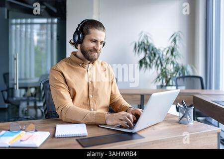 A smiling man wearing headphones works on a laptop at his office desk, focused on his work. The office appears modern. Stock Photo