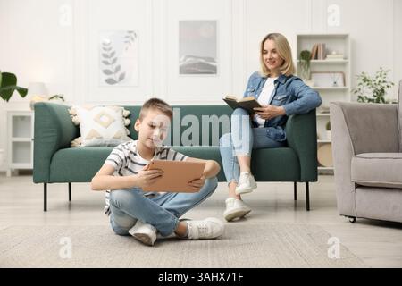 Son using tablet while his mother reading book at home, selective focus Stock Photo