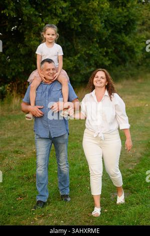 Cute family is walking together in park on summer day. Happy dad is carrying daughter on neck, and mom is walking next to him. Parents with child are spending time outside city, walking in nature Stock Photo