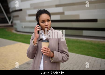 Serious African American woman in coat standing on city street, talking on cell phone, holding cup of coffee in other hand. Concept people in city. Stock Photo
