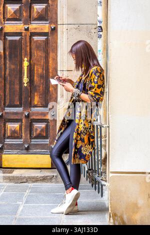 Young modern well-dressed Asian woman outside doorway looking at her smartphone - Paris, France. Stock Photo