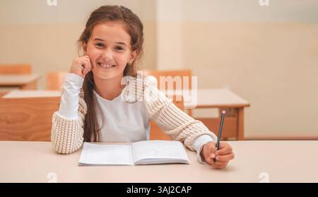 A happy kid girl sitting at a school desk in the classroom, looking at the camera and smiling.  Stock Photo