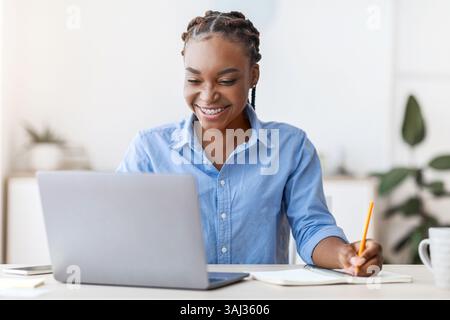 Online Education. Young Black Female Student Using Laptop At Home, Taking Notes Stock Photo