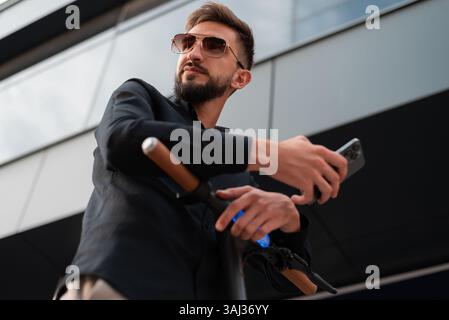 A young happy hipster man with an electric scooter standing near the urban city street and using a mobile phone. Stock Photo