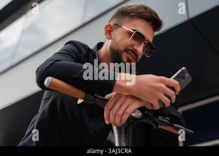 A young happy hipster man with an electric scooter standing on the city street and using a mobile phone. Stock Photo