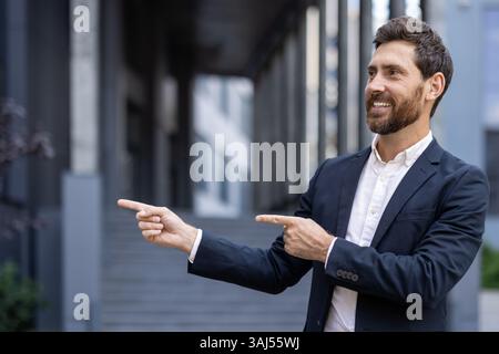A smiling businessman in a dark suit points both fingers to the right, near an office building, ready to lead. Stock Photo