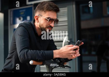 A young hipster man with an electric scooter standing in front of a modern business building on the city street and using a mobile phone. Stock Photo