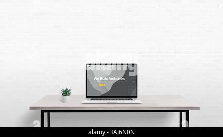 Simple work desk with laptop and plant, conceptual web design displayed on screen, white brick wall in background with empty space for promotional cop Stock Photo