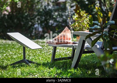 Paris, France - Apr 9, 2025: A Starlink satellite antenna stands beside a lounge chair in a sunny garden with a stack of newspapers resting on the chair' s side table in a relaxed and connected setting Stock Photo