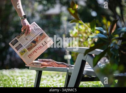 Paris, France - Apr 9, 2025: A hand wearing a smartwatch places a copy of FT Weekend with the headline on Russia sanctions onto a stack of newspapers beside a lounge chair in a peaceful garden Stock Photo