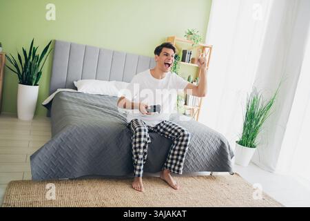 Young man enjoying leisure at home in pajamas while playing video games in a bright bedroom with comfortable decor and natural light Stock Photo