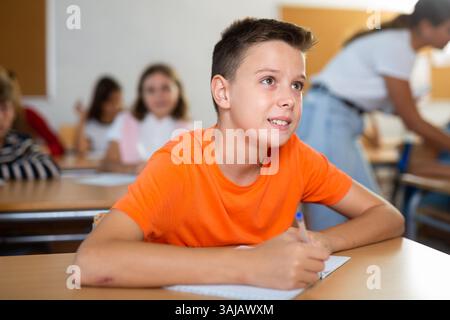 Tween boy listening to schoolteacher and writing in notebook in classroom Stock Photo
