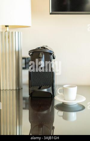 A modern coffee machine is placed on a polished table, accompanied by a small white cup on a saucer. Soft light from a nearby lamp creates a cozy atmo Stock Photo