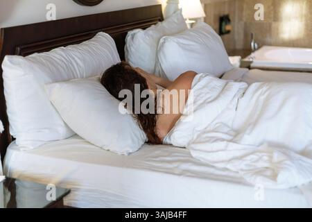 A woman lies on her side in a comfortable hotel room, her face hidden by pillow, enjoying a peaceful moment in soft, bright bedding on a serene mornin Stock Photo
