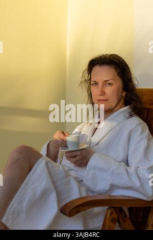 A woman sits comfortably in a wooden chair, dressed in a white robe while sipping coffee. Sunlight softly illuminates the tranquil space, creating a p Stock Photo