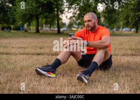 A young sporty man relaxing after exercise while using a phone in the park. Stock Photo