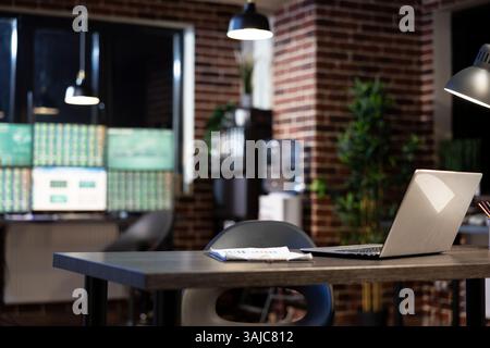 A vacant desk in modern office with a laptop and clipboard, set against a brick wall at night. Blurred computer monitors in the background create a professional and minimalist workspace. Stock Photo