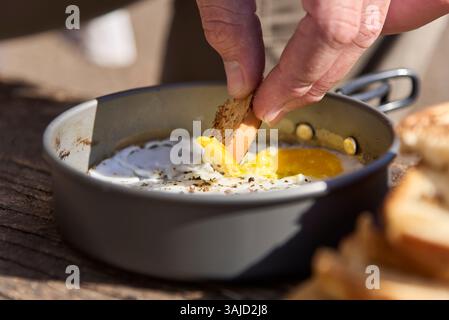 Close up view of a hand dipping a piece of toasted bread into the yolk of a fried egg cooked in a camping pan. Stock Photo