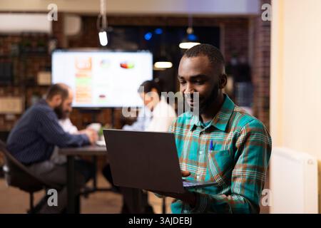 Professional black man deeply engaged in project planning, reviews financial analytics on his laptop. Behind him, coworkers discuss business trends, creating a dynamic workplace scene. Stock Photo