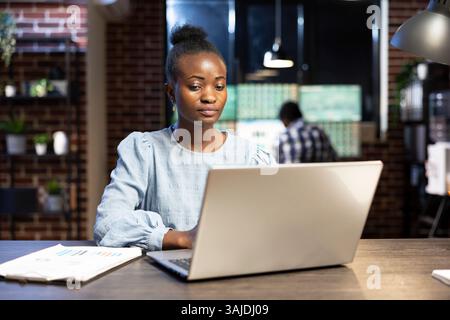 Determined female entrepreneur analyzing financial data on laptop at her desk, surrounded by trading documents. Her coworker, slightly blurred, works in the background at a multiscreen setup. Stock Photo