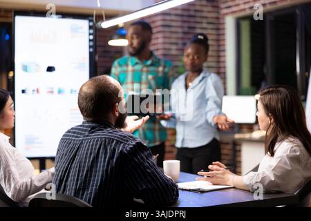 Multiethnic coworkers work efficiently in tracking progress on financial report and completing project plans. African american employees doing business presentation in late night group meeting. Stock Photo