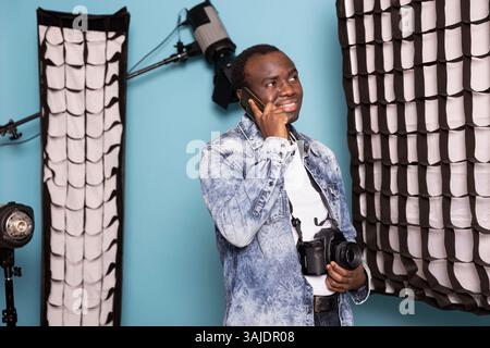 Young black man in a denim jacket, with a DSLR camera hanging around his neck, talking on his mobile phone. The smiling male photographer stands confidently in a studio, engaged in conversation. Stock Photo