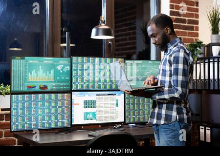 Male financial analyst with a laptop, engaged in forex trading at night, reviewing currency exchange charts. Black man standing and analyzing investment data on multiple screens in modern workstation. Stock Photo