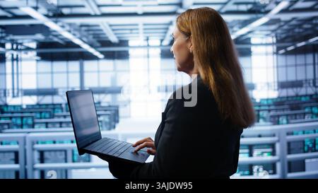 System administrator in server hub checking recovery plan on laptop, monitoring energy consumption. IT expert uses notebook in data center workspace, ensuring rigs are functioning optimally, camera B Stock Photo