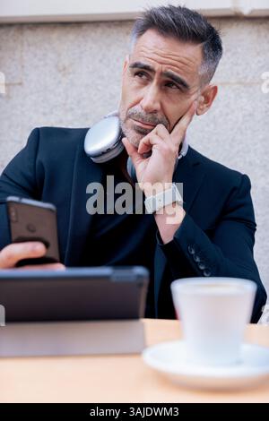 Man with headphones thinking while using phone and tablet at outdoor cafe table Stock Photo