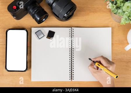 A person is writing on a tablet with a pen. a phone with a screen mockup, a coffee cup of writing supplies, a pens, a notepad on a white wooden table Stock Photo
