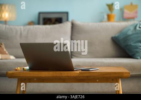Close-up of open laptop on wooden desk in cozy living room setting surrounded by smartphone, tablet, pen, lamp, and bookshelf in background Stock Photo