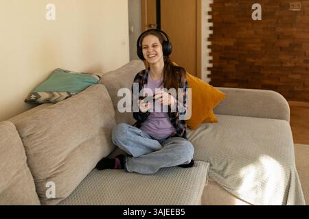 Young woman enjoys gaming while wearing headphones in cozy living room setting during afternoon Stock Photo